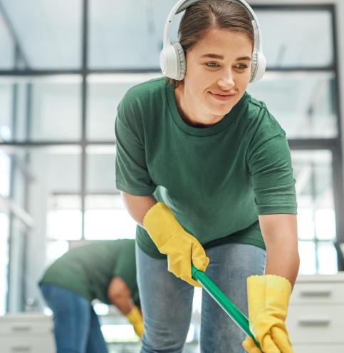 Shot of a young woman cleaning an office with her colleague in the background.